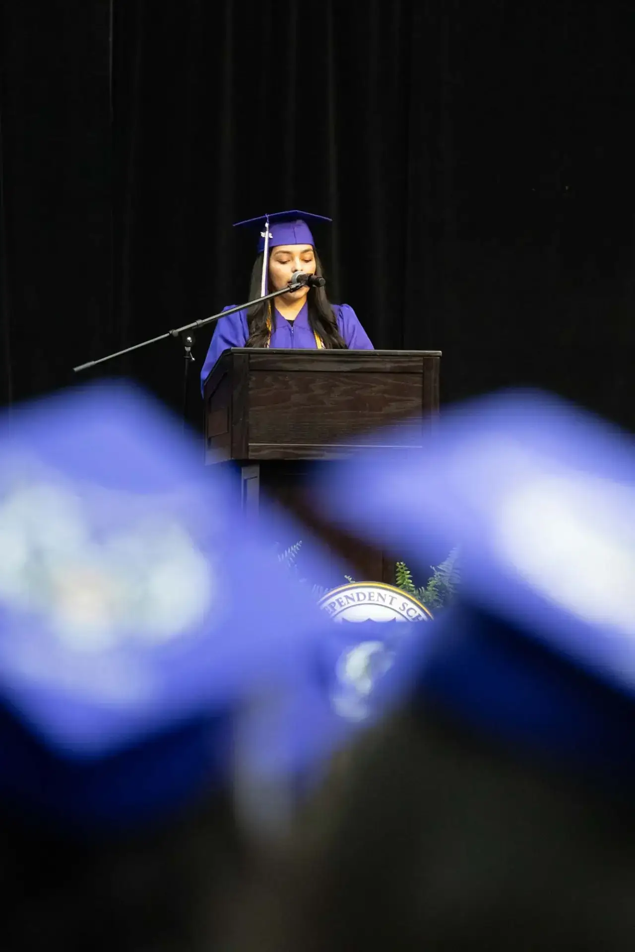 Una mujer con toga y birrete de graduación púrpura está de pie en un podio, hablando por un micrófono. Los birretes azules de los graduados sentados se difuminan en primer plano. El ambiente es formal y festivo.