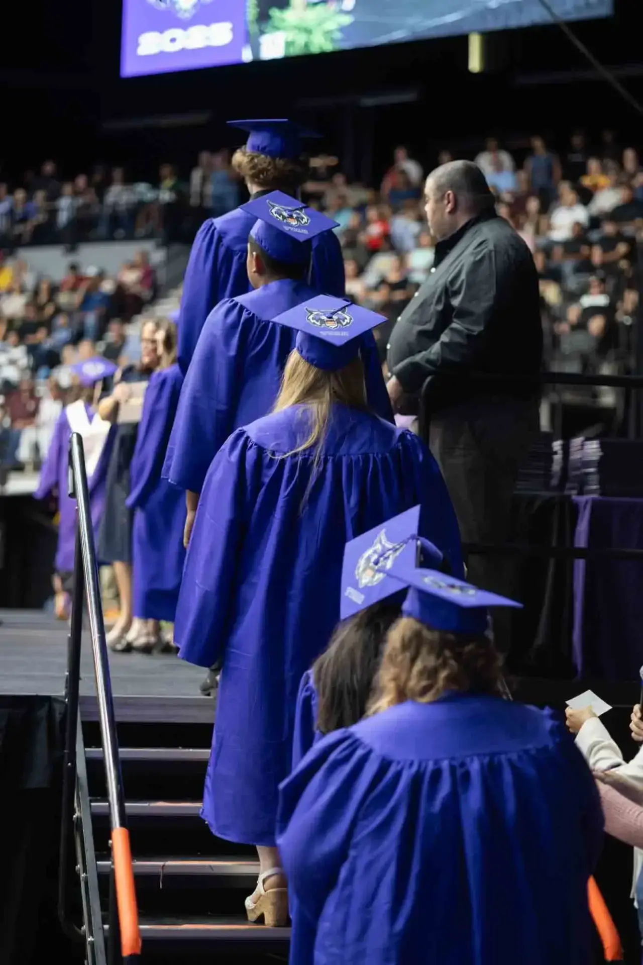 Graduados con togas azules suben al escenario en una ceremonia, llevando birretes decorados. Una multitud observa al fondo, creando un ambiente de celebración.