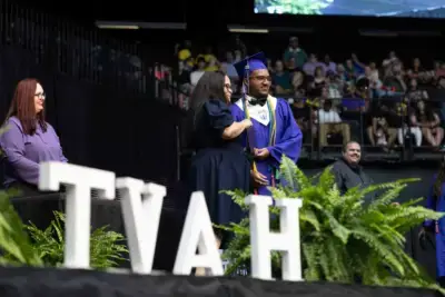 Graduada con toga y birrete recibe un diploma en el escenario de una ceremonia, sonriendo con el profesorado. Letras "TVAH" y helechos verdes adornan el primer plano.