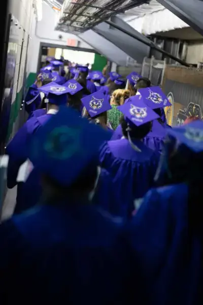 Graduados con togas moradas y birretes caminan por un pasillo, con la expectación en el aire. Las gorras presentan diseños personalizados, añadiendo un tono festivo.