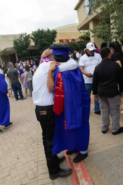 Una graduada con toga y birrete azules abraza a un adulto mayor vestido de blanco en el exterior de un local. La escena es alegre, con una multitud celebrando en el fondo.