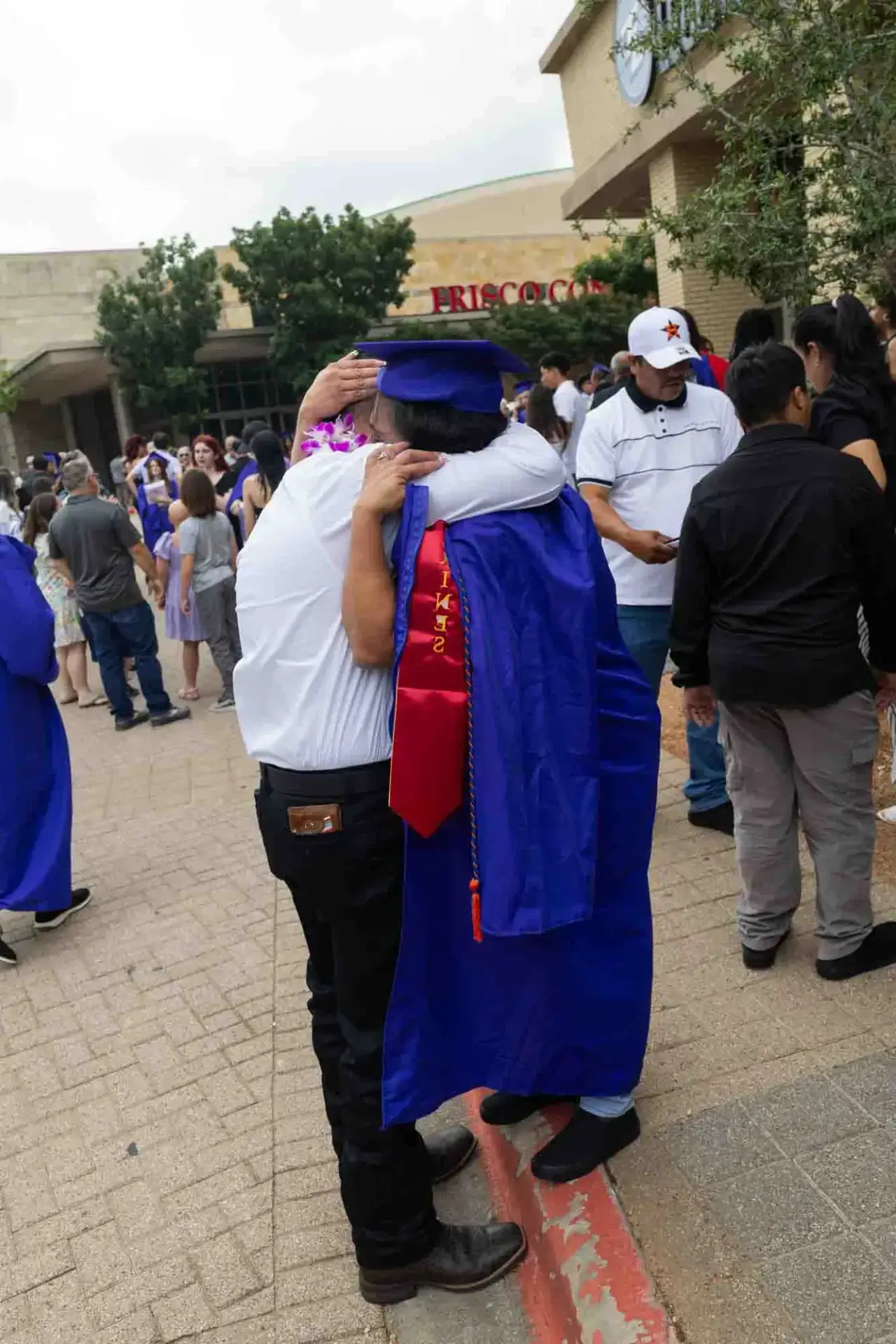 Una graduada con toga y birrete azules abraza a un adulto mayor vestido de blanco en el exterior de un local. La escena es alegre, con una multitud celebrando en el fondo.