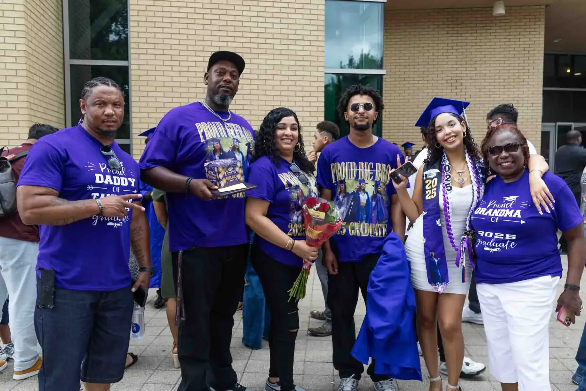 Una familia alegre celebrando la graduación. Seis personas permanecen juntas fuera, llevando camisetas con mensajes de apoyo. El graduado, con toga y birrete, sonríe alegremente.