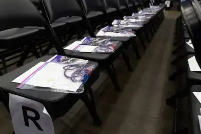 Filas de sillas negras con paquetes en cada asiento, que incluyen un folleto y un cordón de colores. Un papel con la etiqueta "R" marca una fila. El entorno es tranquilo y organizado.