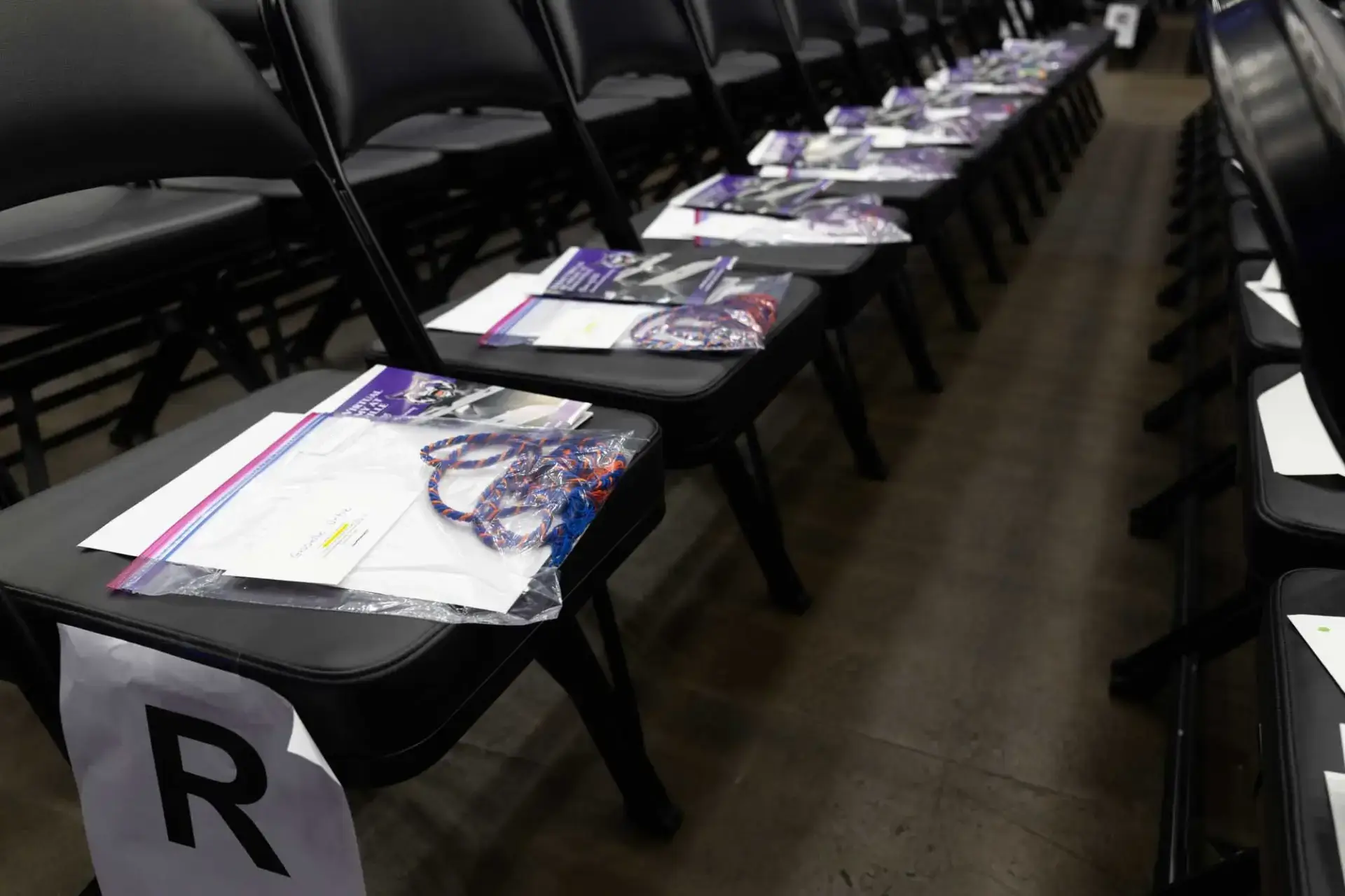 Filas de sillas negras con paquetes en cada asiento, que incluyen un folleto y un cordón de colores. Un papel con la etiqueta "R" marca una fila. El entorno es tranquilo y organizado.