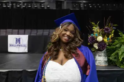 Graduada sonriente con toga y birrete azul de pie en el escenario con la pancarta de Hallsville al fondo. Un ramo de flores de colores está cerca, transmitiendo alegría.