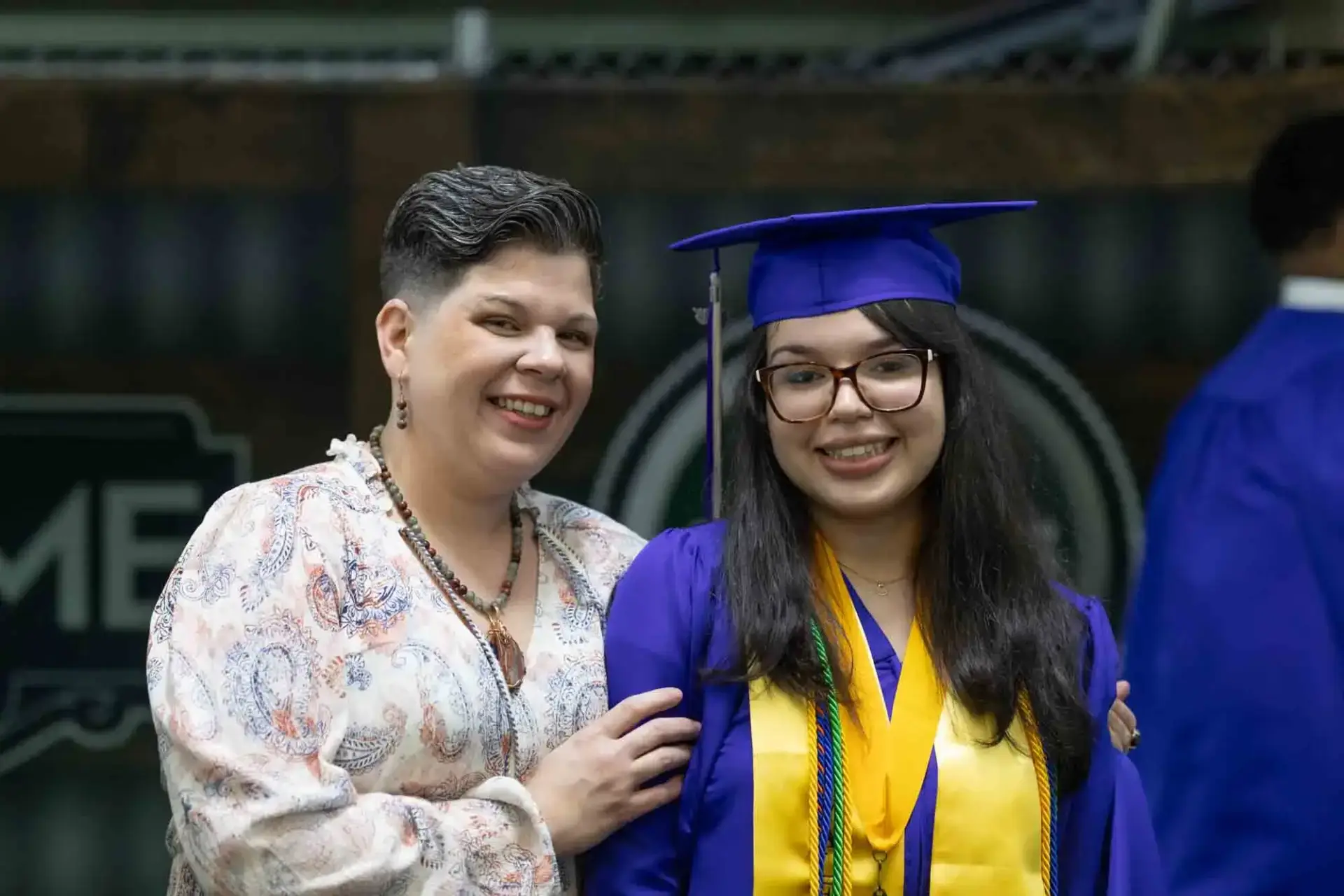 Un graduado sonriente con toga y birrete azules está de pie junto a una mujer orgullosa con el pelo corto. La escena transmite alegría y logro.