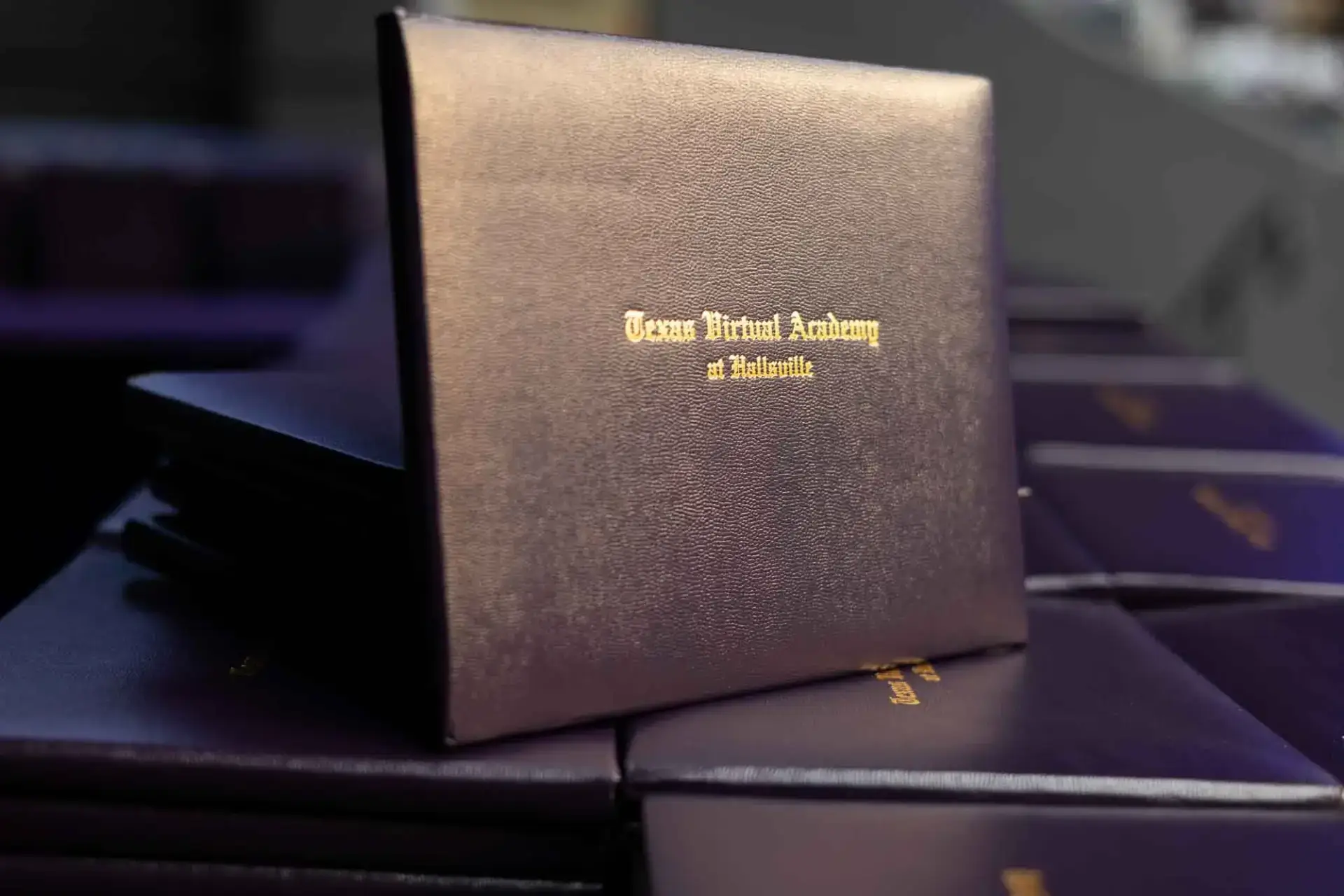 Pila de fundas oscuras para diplomas con texto dorado en el que se leeTexas Virtual Academy at Hallsville", símbolo de graduación y logro.