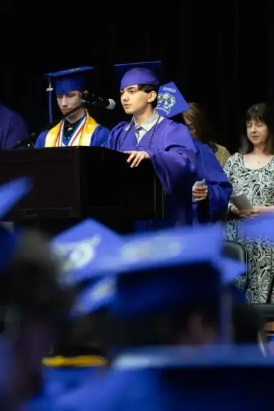 Un graduado con toga y birrete azules habla en un podio sobre el escenario durante una ceremonia. Un compañero se sitúa a su lado y la gente observa al fondo.