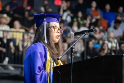 Un graduado con toga y birrete azules habla con confianza en un podio, dirigiéndose al público. El fondo muestra una multitud borrosa entre los asistentes.