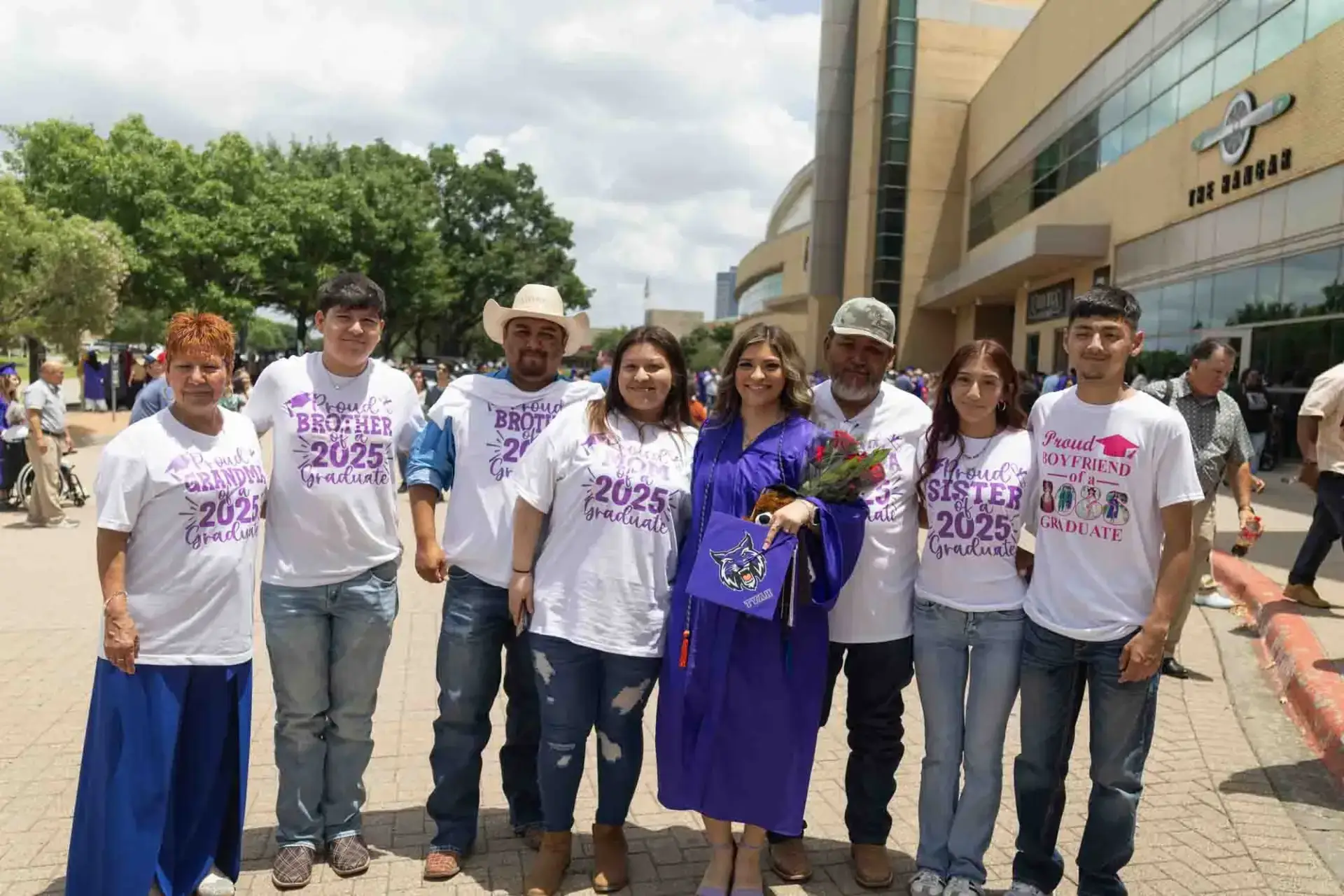 Un grupo de ocho personas posa al aire libre. Una mujer con un vestido de graduación morado sostiene unas flores, rodeada de familiares que la apoyan y que llevan camisetas personalizadas. El ambiente es de celebración.
