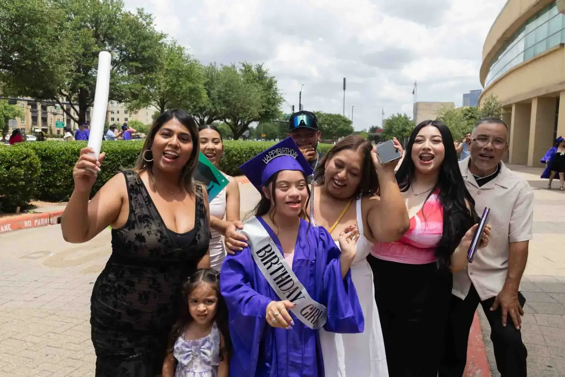 Feliz graduada con toga y birrete púrpura, llevando un fajín de "Cumpleañera", rodeada de alegres familiares que sostienen un diploma. Celebración al aire libre, día soleado.