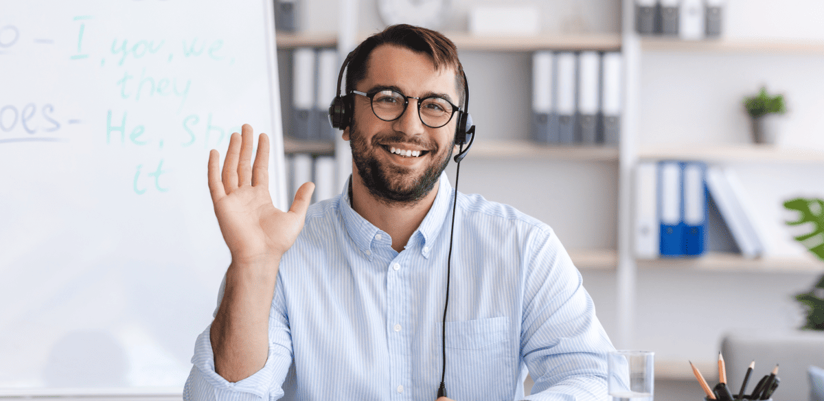 Un hombre sonriente, con camisa azul y gafas, sentado ante un escritorio, levanta las manos en un gesto de excitación o celebración.
